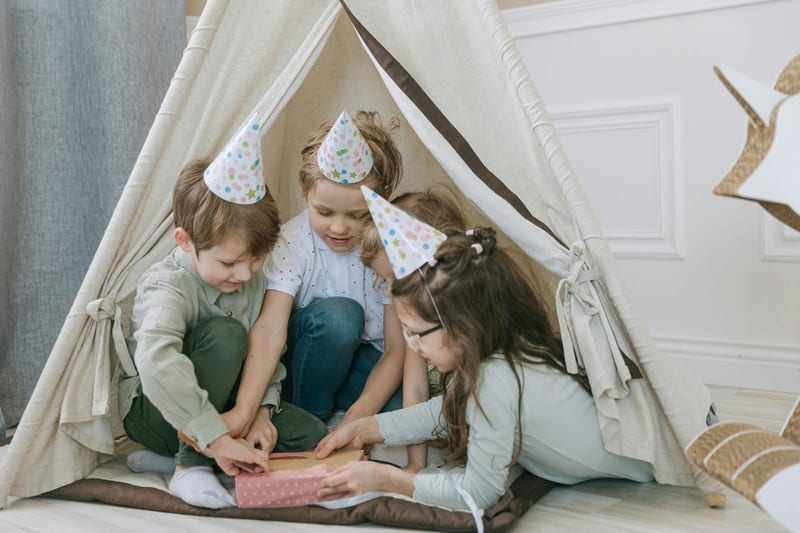 Kids wearing party hats open a gift inside a teepee, celebrating a birthday indoors.
