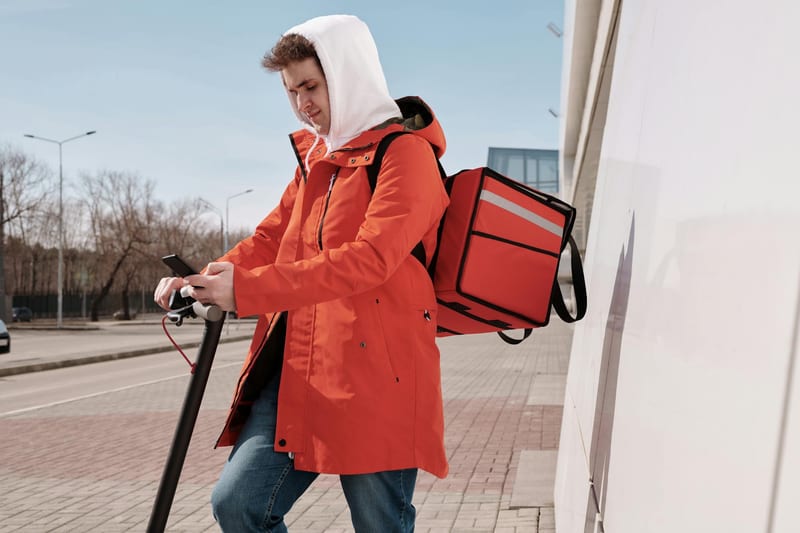 Man in orange jacket using an electric scooter for food delivery in a city street, checking his smartphone.