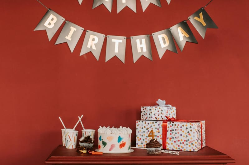 Festive birthday scene with cake, cupcakes, gifts, and decorations against red wall.